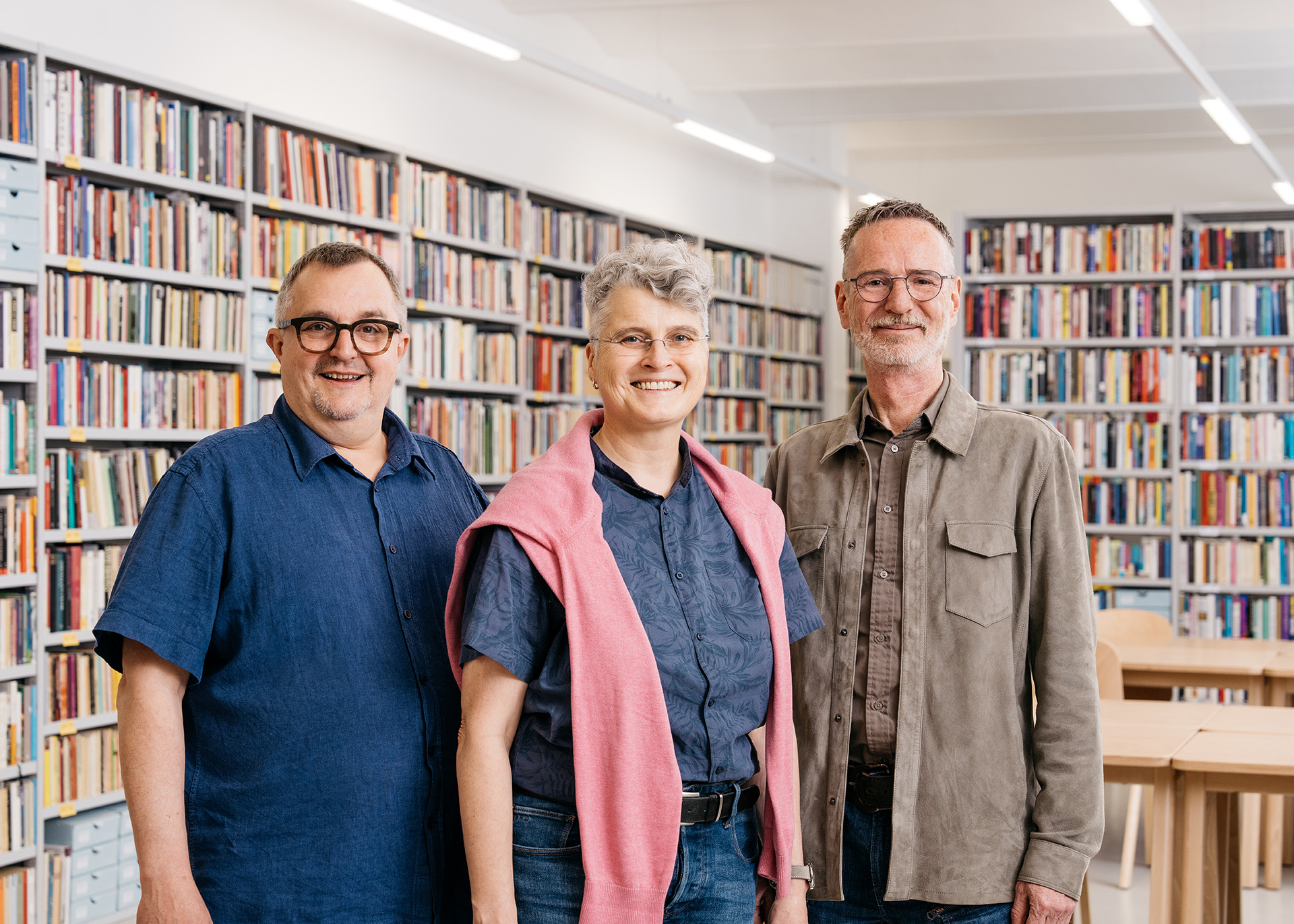 Drei Personen lächeln in einer Bibliothek vor Regalen voller Bücher.