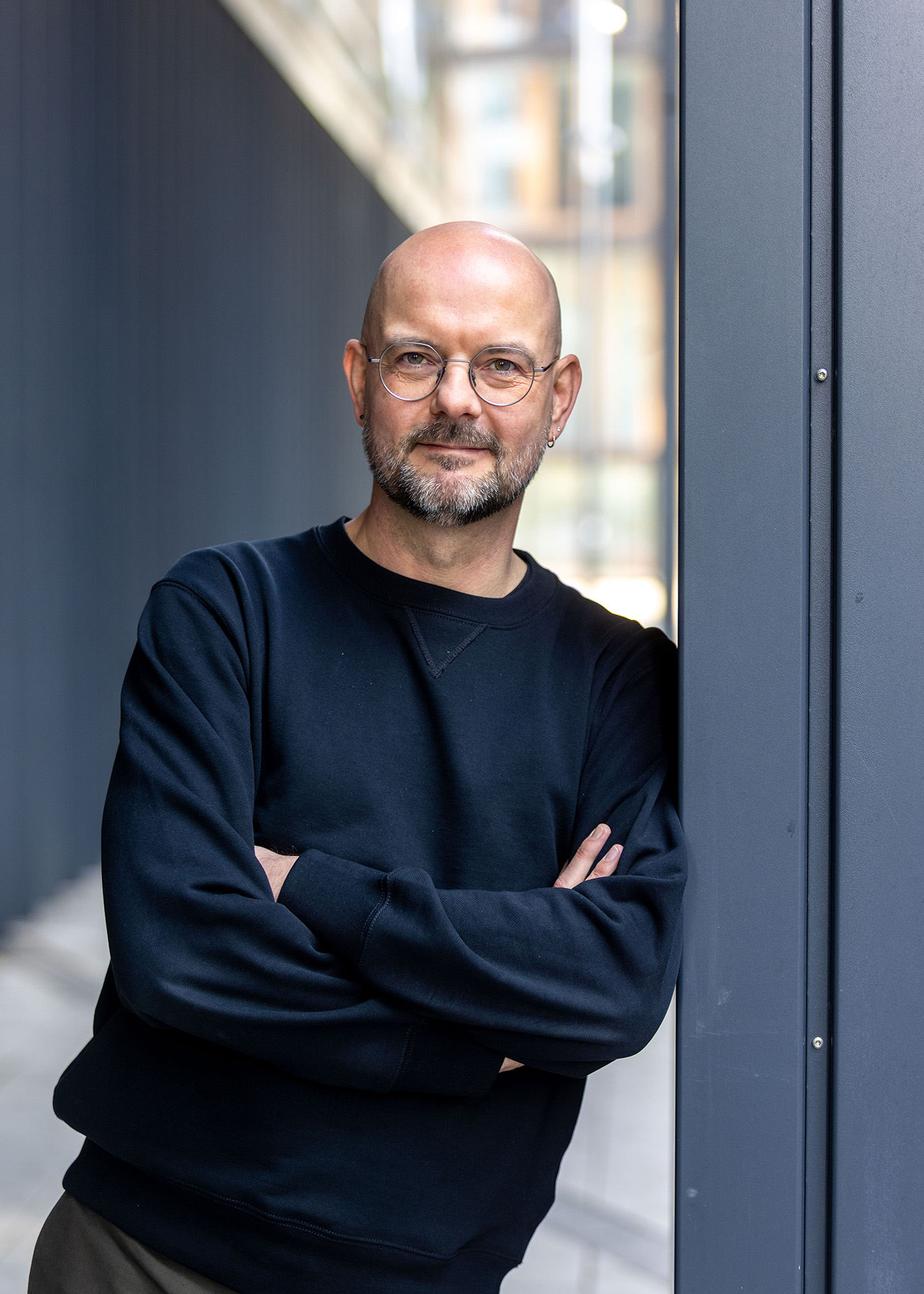 Person mit Brille und Bart lehnt lässig an einer Wand und schaut in die Kamera.
