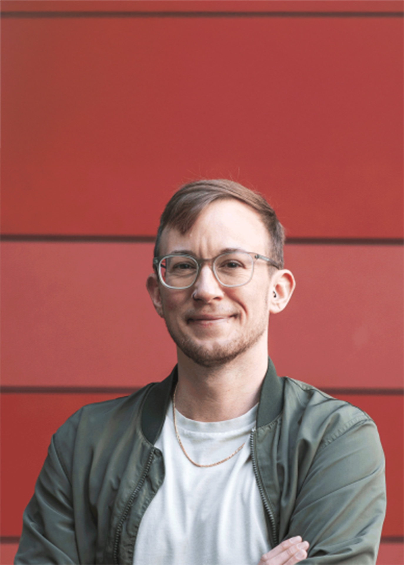 Jonah lächelt vor einer roten Wand, trägt eine grüne Jacke und Brille.