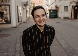 Lächelnde Person in gestreiftem Hemd steht in einer ruhigen Stadtstraße.