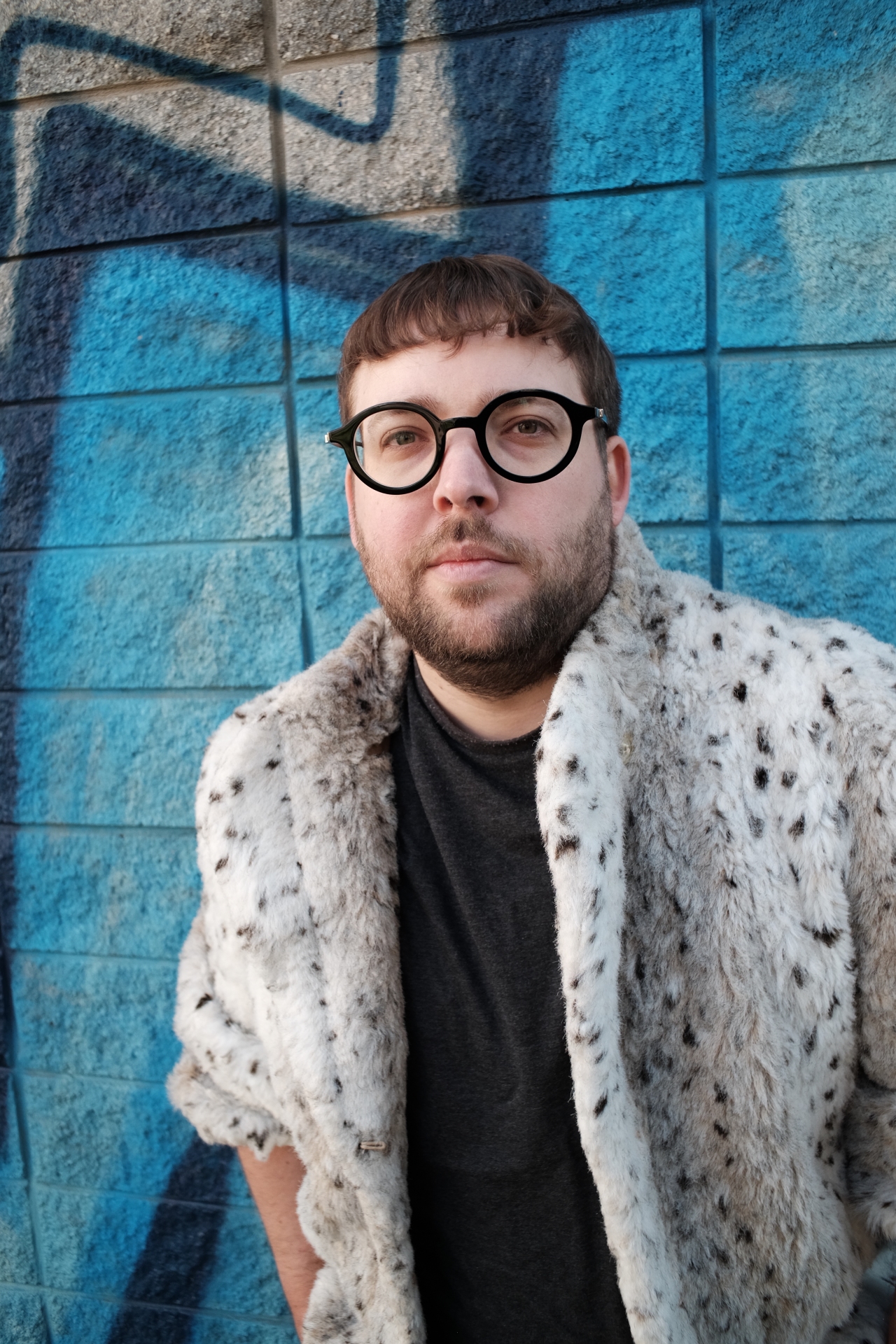 Person in einem auffälligen Pelzmantel vor einer blauen Graffiti-Wand, schaut direkt in die Kamera.