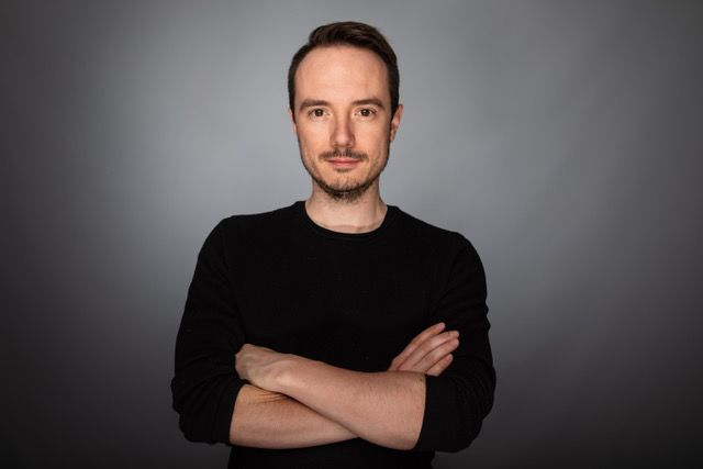 Person mit verschränkten Armen blickt direkt in die Kamera vor einem grauen Hintergrund.