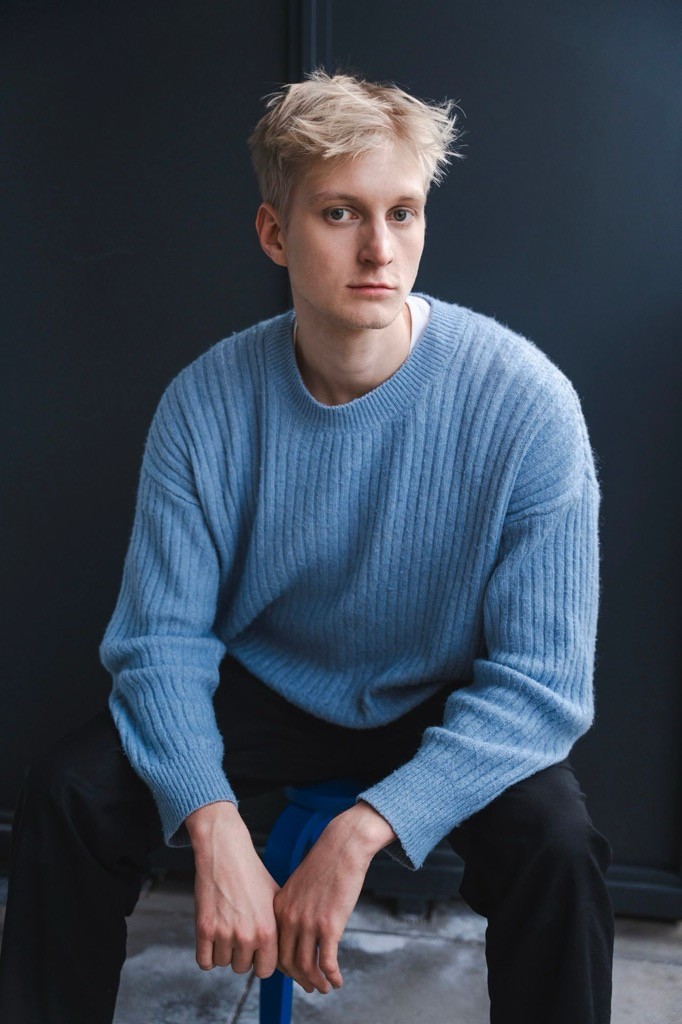 Junger Mensch sitzt auf einem Stuhl, schaut nachdenklich in die Kamera vor dunklem Hintergrund.