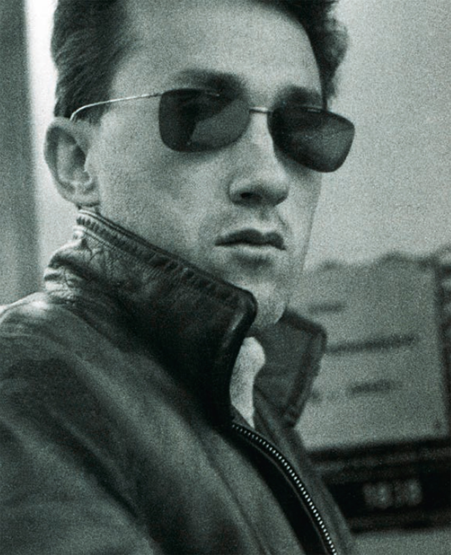 Black-and-white portrait of a man in sunglasses, wearing a leather jacket, looking intently.